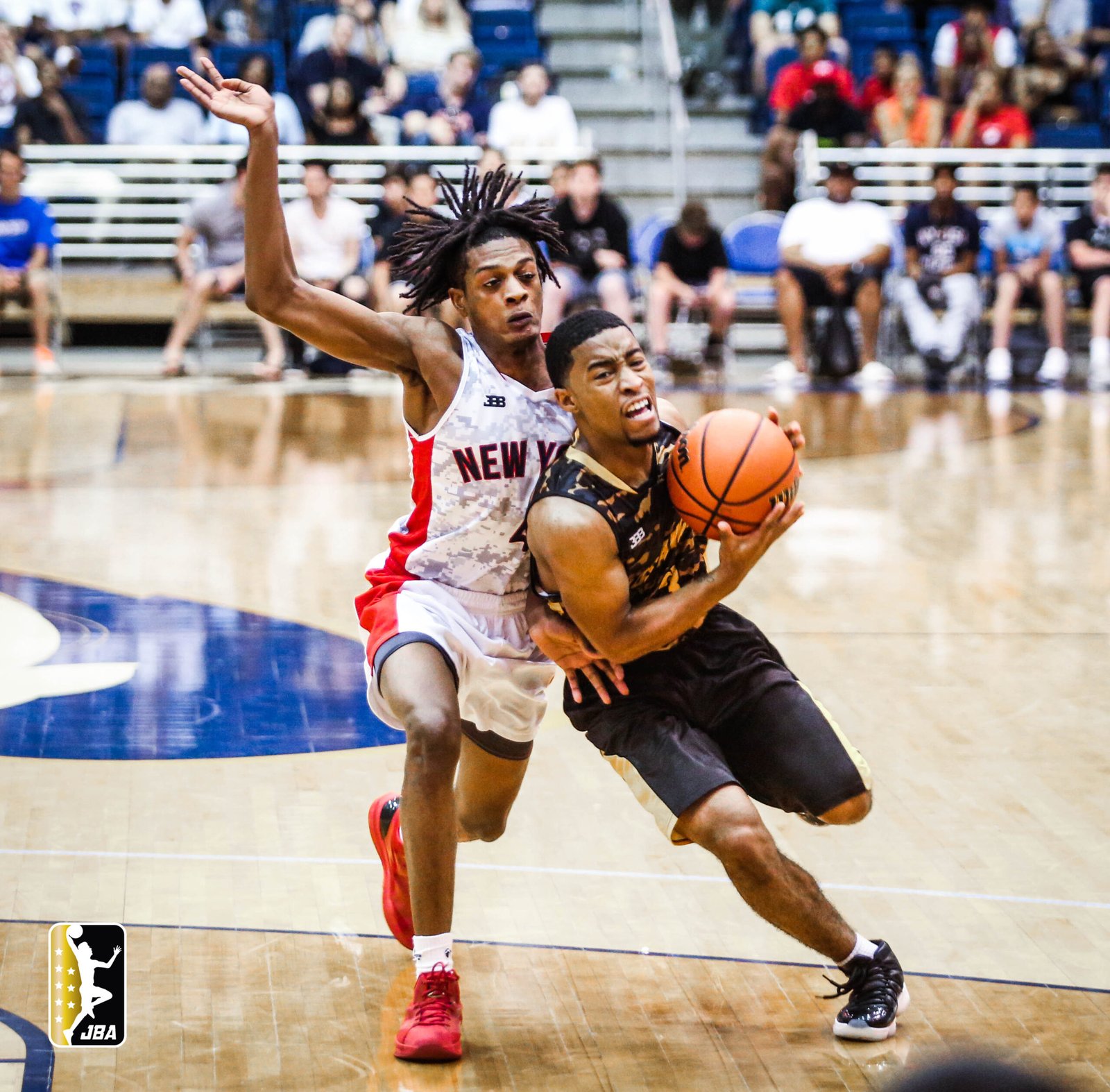 An intense moment on the basketball court during a Junior Basketball Association game, featuring Jameer Killing in a white and red uniform labeled 'NEW YORK' on defense, with his arm outstretched, and an opposing player Niles Malone in a black uniform driving towards the basket with the ball, his face showing determination.