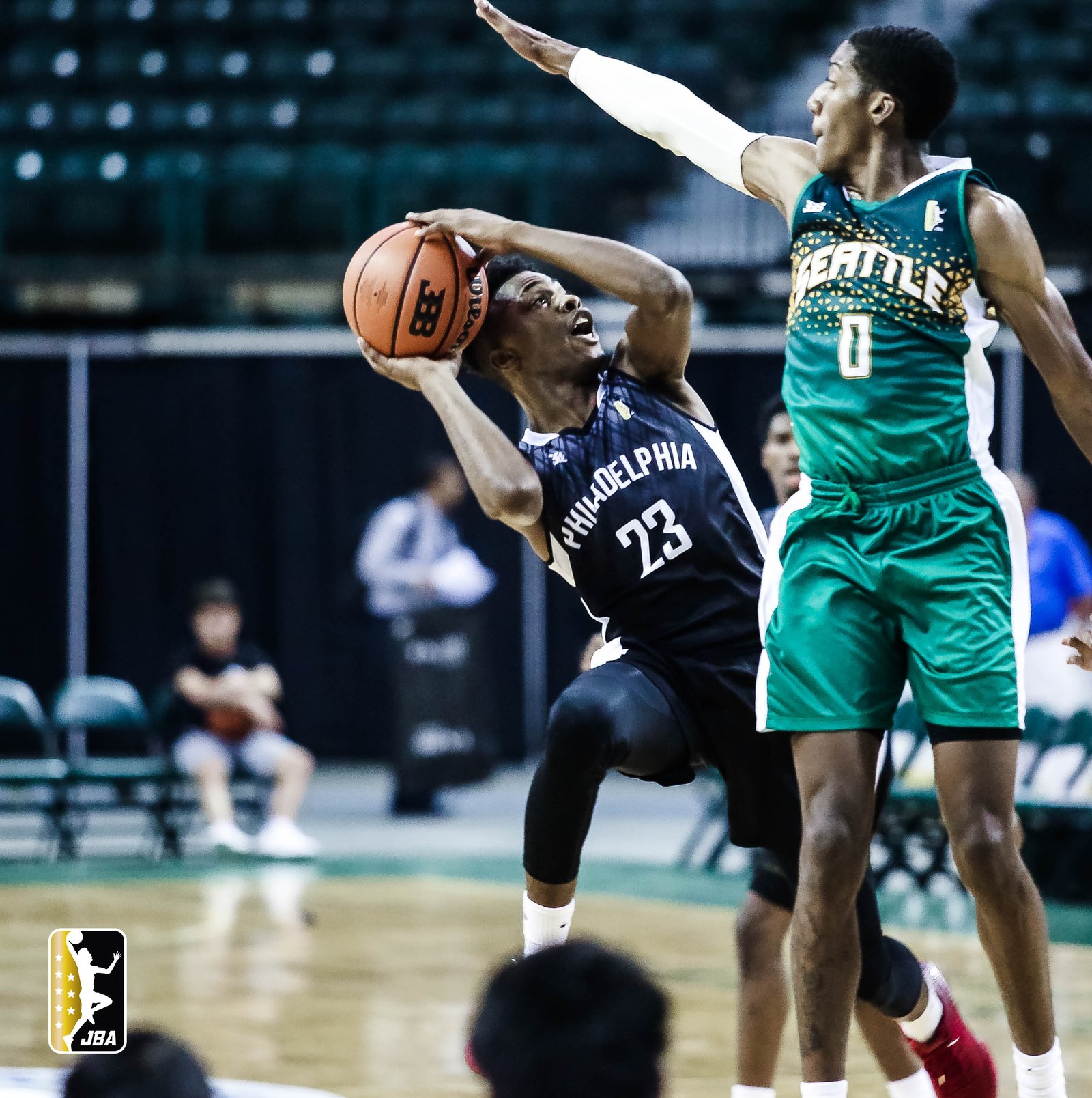 "Jaylen Nixon, wearing a black Philadelphia jersey with the number 23, is in mid-air making an athletic layup attempt against Jerrel Springer, who is in a green Seattle jersey with the number 0, playing defense with his hand up to block the shot during a Junior Basketball Association game.