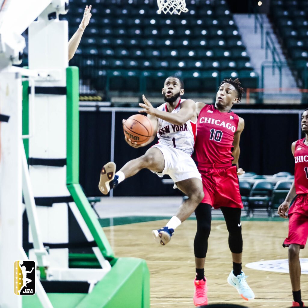 In a dynamic basketball scene, Nolan Irby, in a white New York jersey with the number 1, is captured mid-leap as he drives towards the basket for a layup. Breydo, in a red Chicago jersey with the number 10, is closely trailing the action, focused and ready to defend, this high-stakes Junior Basketball Association game moment